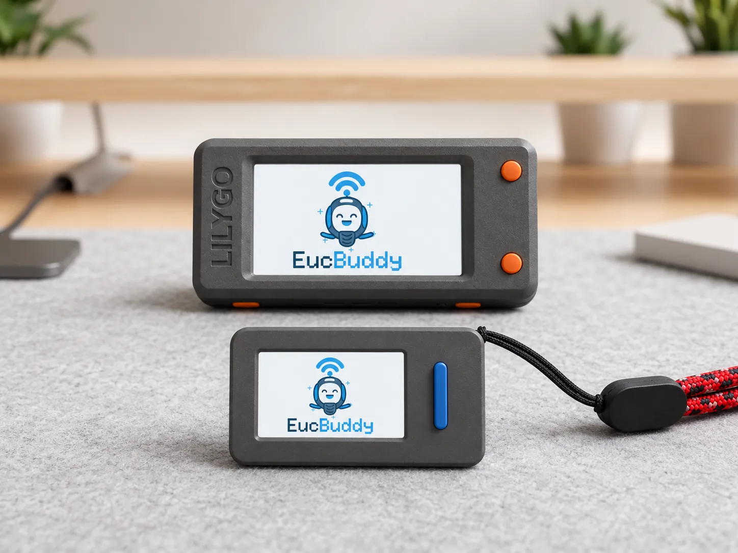Two EucBuddy devices on a desk: a LilyGO T-Display-S3 and a compact M5Stick-S3 on a lanyard, both showing the EucBuddy splash screen with the BLE-waved mascot